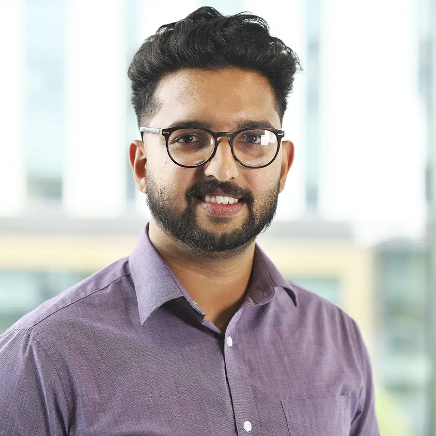 A man with dark hair and a beard, wearing glasses and a light purple button-up shirt, smiles at the camera in a bright indoor setting with blurred windows in the background.