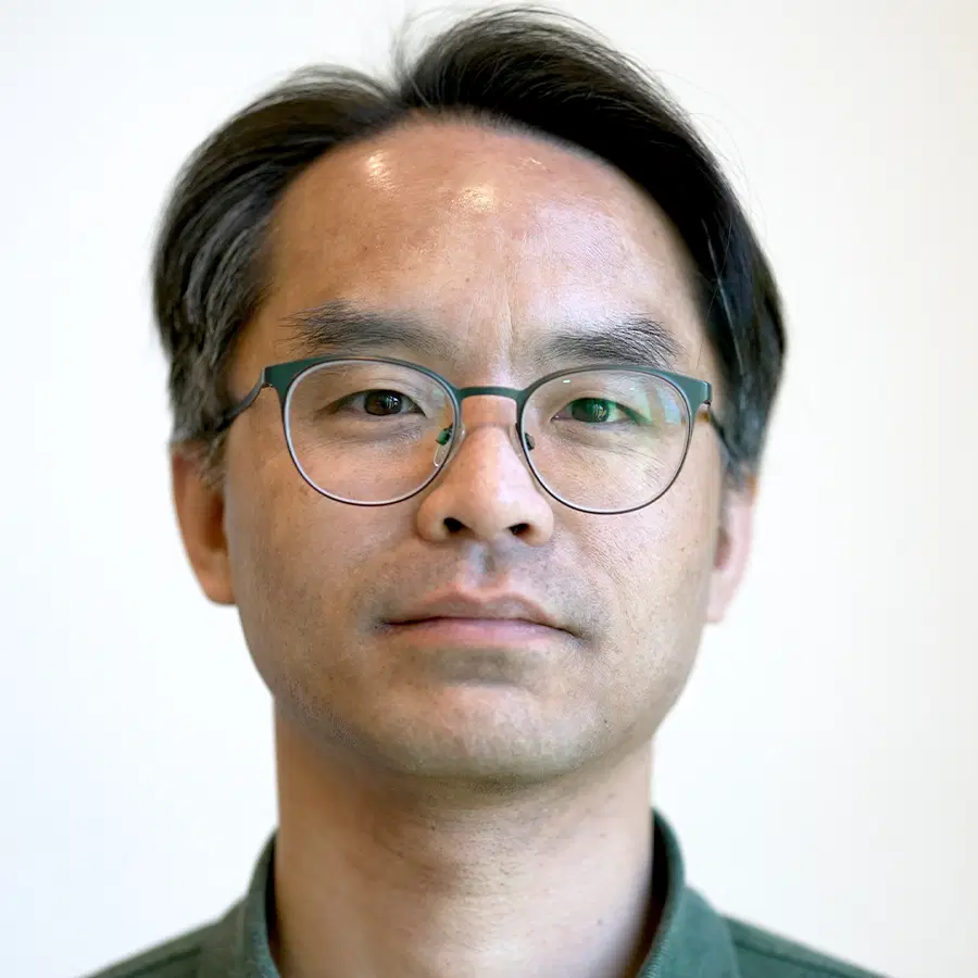 A man with short dark hair and glasses, wearing a grey collared shirt, looks directly at the camera against a plain light background.