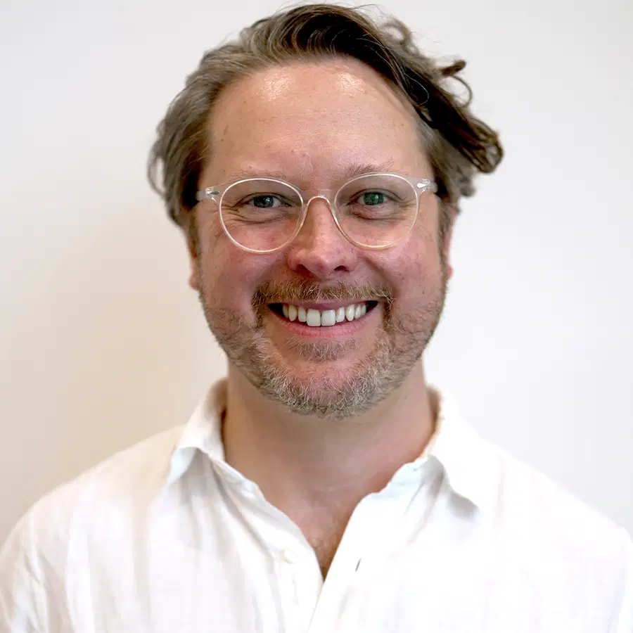 A smiling man with short, wavy brown hair, a beard, and glasses, wearing a white button-up shirt, stands in front of a plain white background.