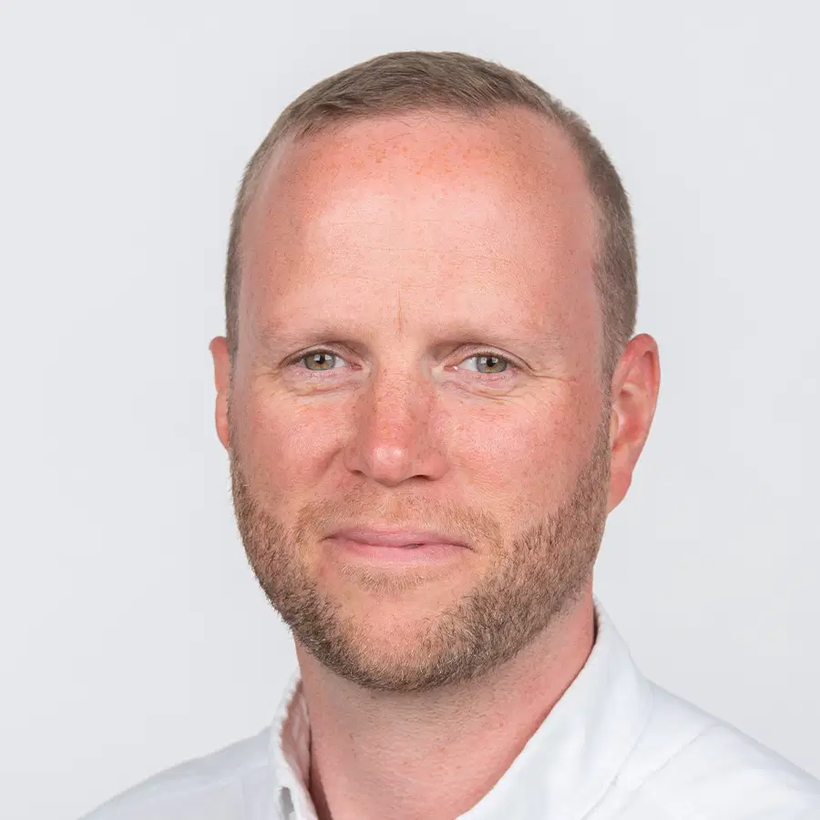 A man with short light brown hair and a trimmed beard, wearing a white collared shirt, poses against a plain light grey background, softly smiling at the camera.