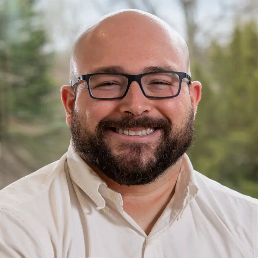 Bald man with glasses and a beard, wearing a white shirt, smiling in front of a window with a blurred, green outdoor background.