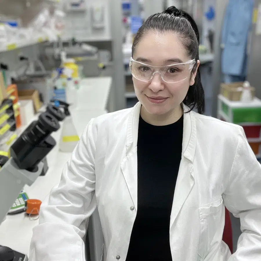A woman wearing safety glasses and a white lab coat stands in a laboratory, smiling at the camera. Lab equipment and supplies are visible on the benches around her.