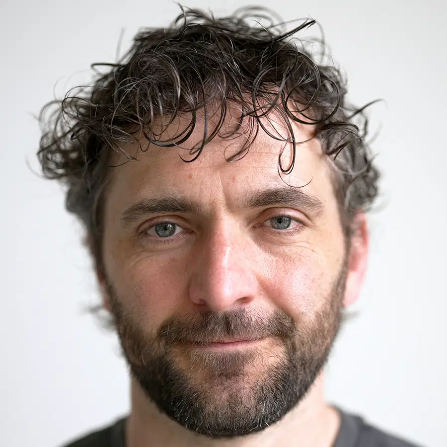 A man with curly brown hair, a short beard, and a neutral expression looks directly at the camera. He is in front of a plain light grey background.