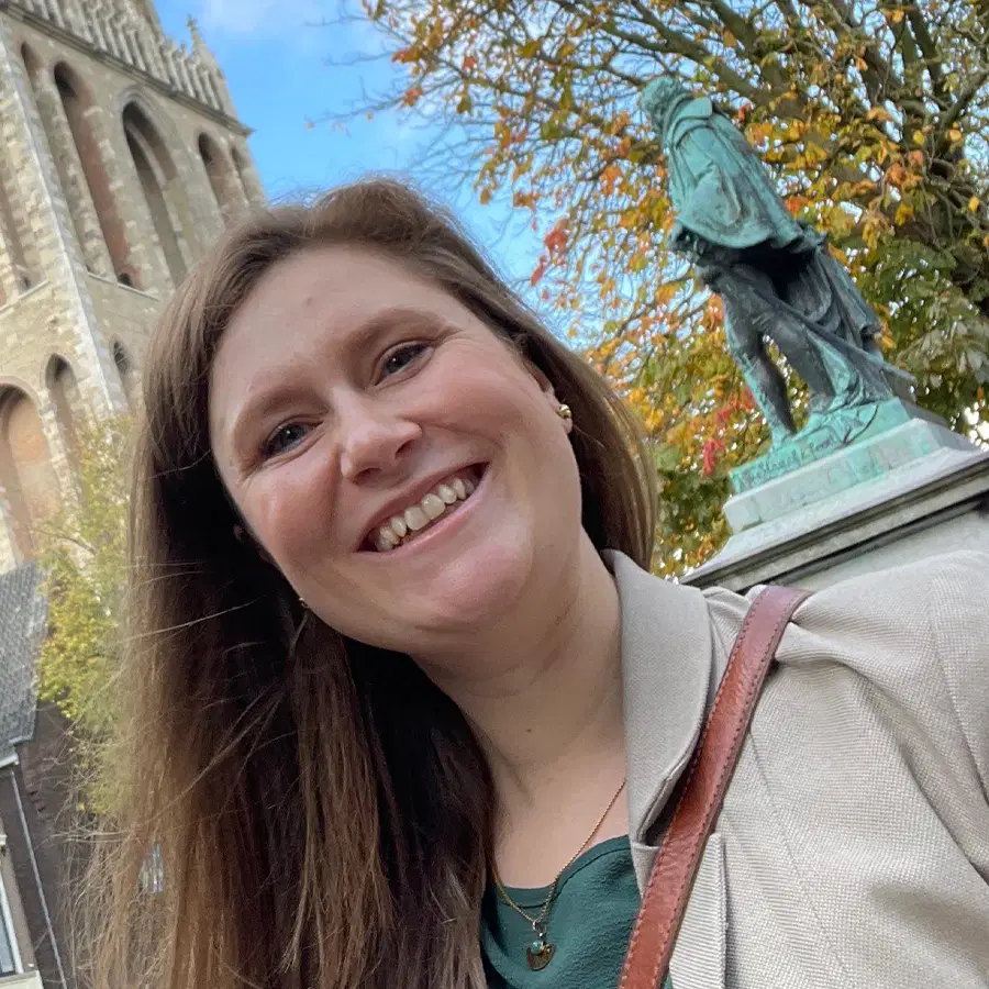 A woman with long brown hair smiles at the camera outdoors near a statue and historic building, with autumn trees and blue sky in the background.