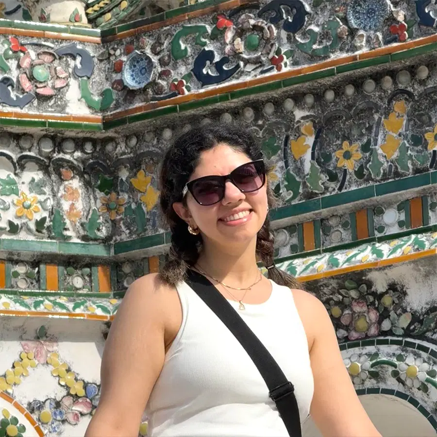 A woman in sunglasses and a white top smiles in front of a colourful, ornate building decorated with floral patterns and tiles.