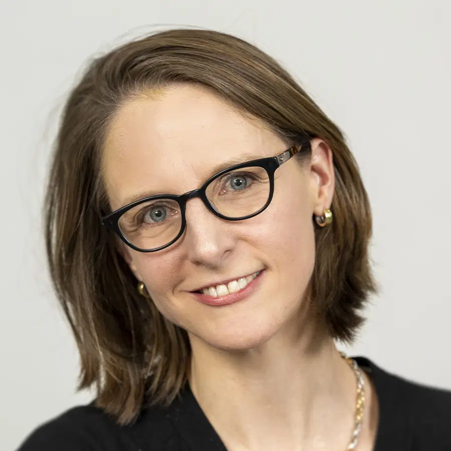 Smiling woman with straight brown hair, wearing black-rimmed glasses, silver earrings, and a necklace, poses in front of a light grey background.
