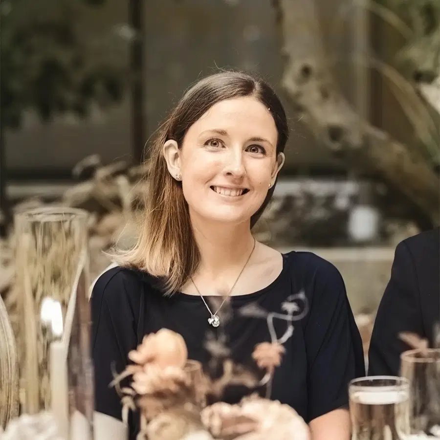 A woman with light brown hair in a navy dress smiles at a dinner table, surrounded by flowers and glasses, with warm lighting and a blurred background.