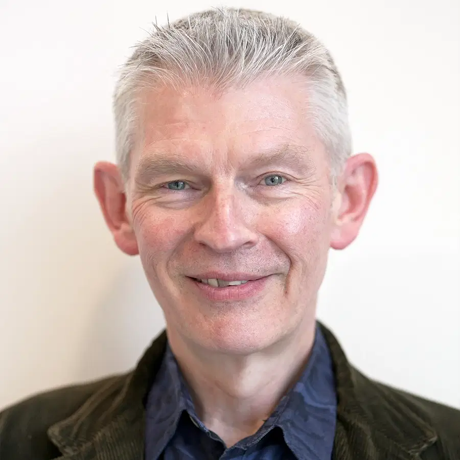 Smiling older man with short grey hair, wearing a dark blazer and a blue shirt, posed against a plain white background.