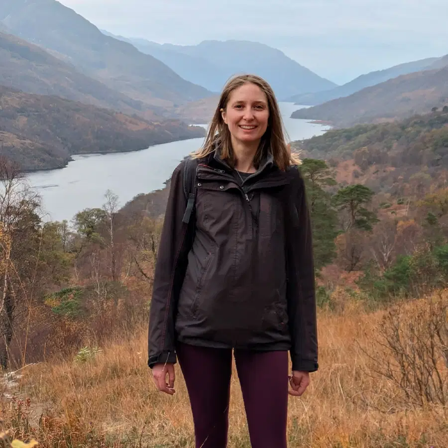 A woman wearing a dark jacket stands smiling in front of a scenic landscape with a river, hills, and mountains under a cloudy sky.