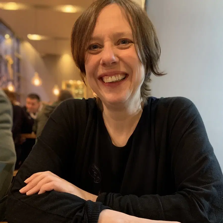 A person with short brown hair, wearing a black shirt, is smiling whilst sitting at a table in a warmly lit indoor setting with blurred people and lights in the background.