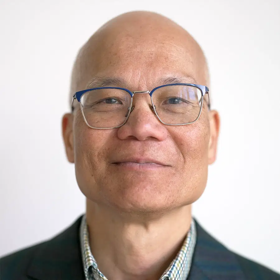 A middle-aged man with glasses, a bald head, and a slight smile, wearing a checked shirt and a dark jacket, standing in front of a plain light background.