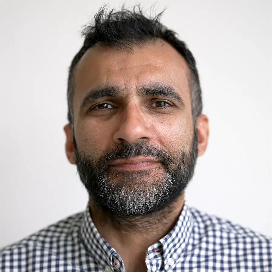 A man with short dark hair and a beard, wearing a blue and white chequered shirt, looks at the camera with a neutral expression against a plain light background.