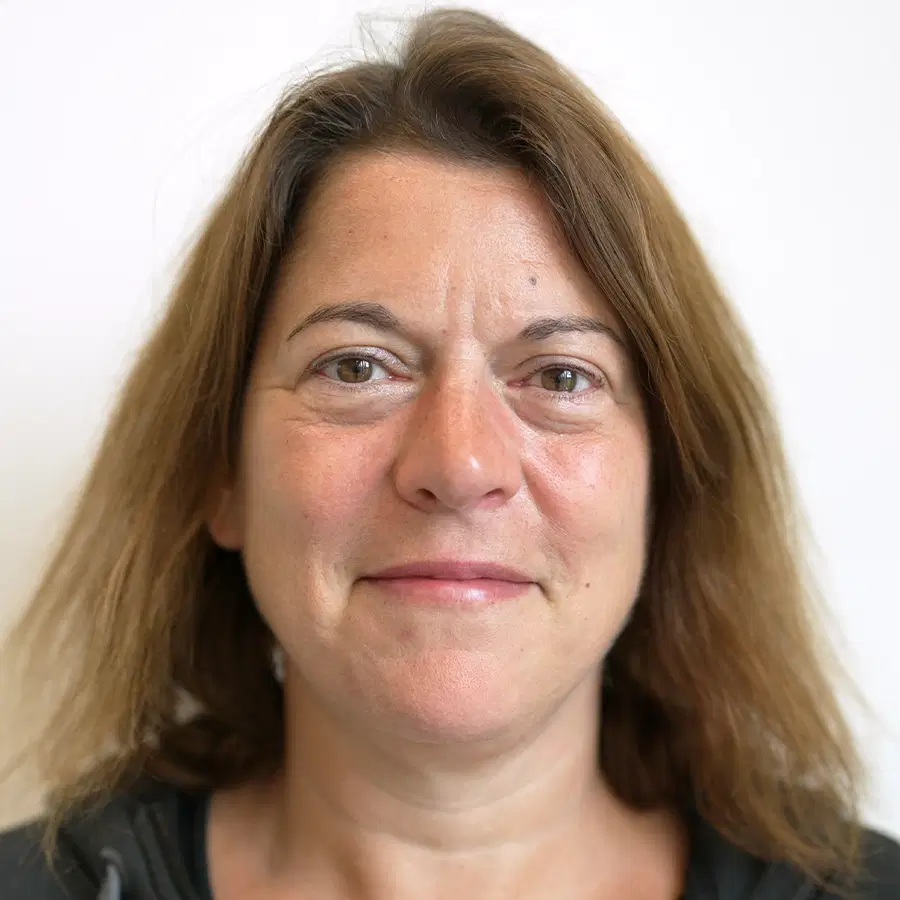 A woman with shoulder-length brown hair, wearing a dark top, smiles softly whilst looking directly at the camera against a plain white background.
