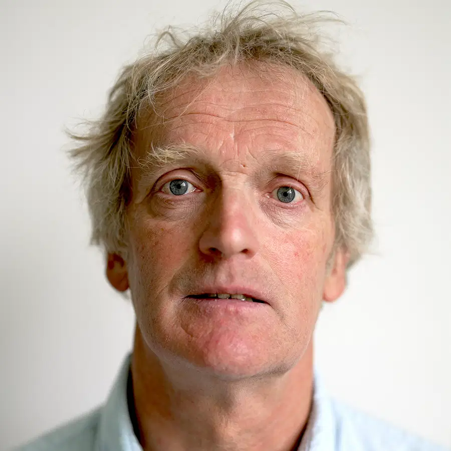 A middle-aged man with light skin, grey messy hair, and a neutral expression looks directly at the camera against a plain white background.