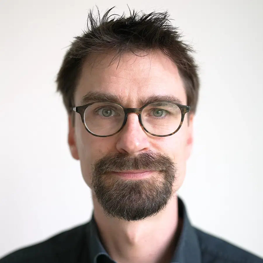 A man with short, dark hair, glasses, and a beard looks directly at the camera, wearing a dark shirt, with a neutral light background.