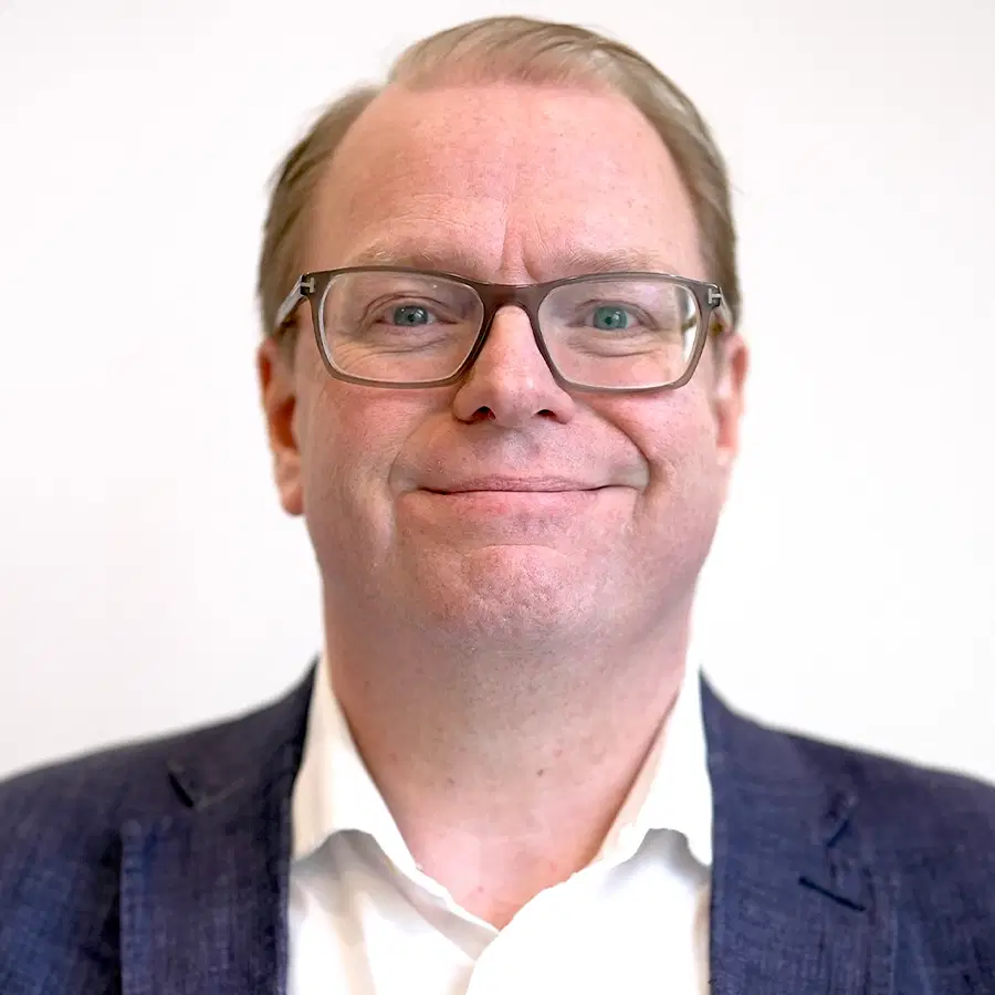 A man with glasses, short fair hair, and a blue blazer over a white collared shirt smiles slightly in front of a plain white background.