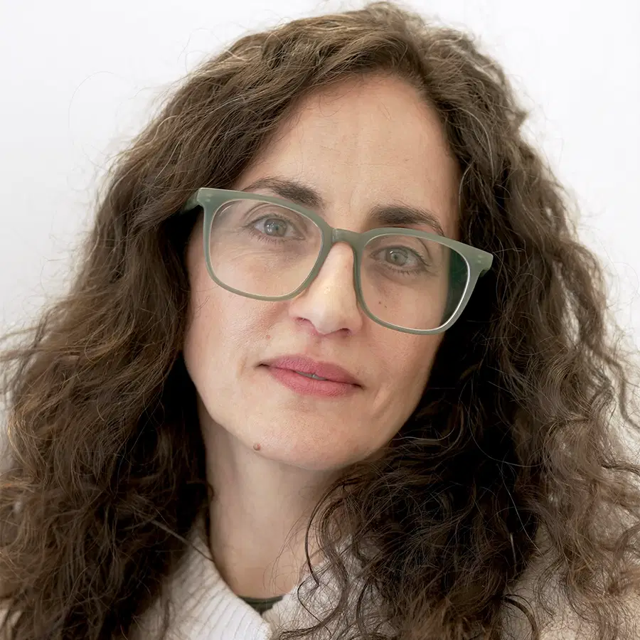 A woman with long curly brown hair, wearing grey glasses and a white jumper, looks at the camera with a neutral expression against a plain light background.