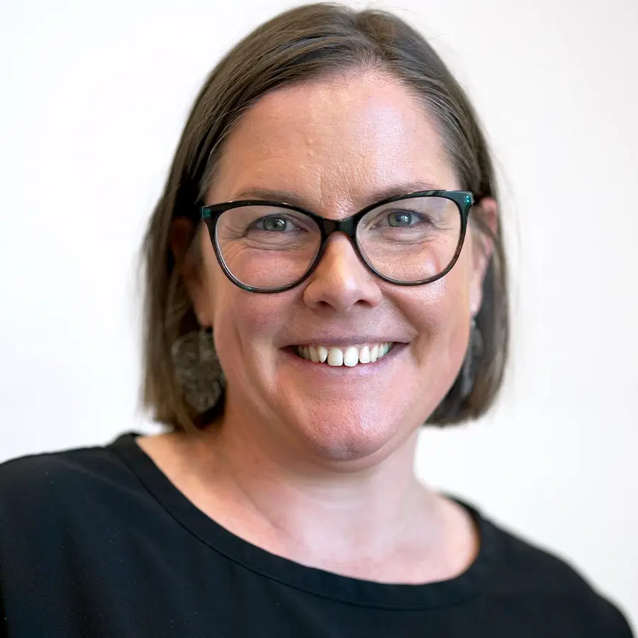 A woman with straight brown hair, glasses, and earrings smiles at the camera. She is wearing a black top and has a light background behind her.