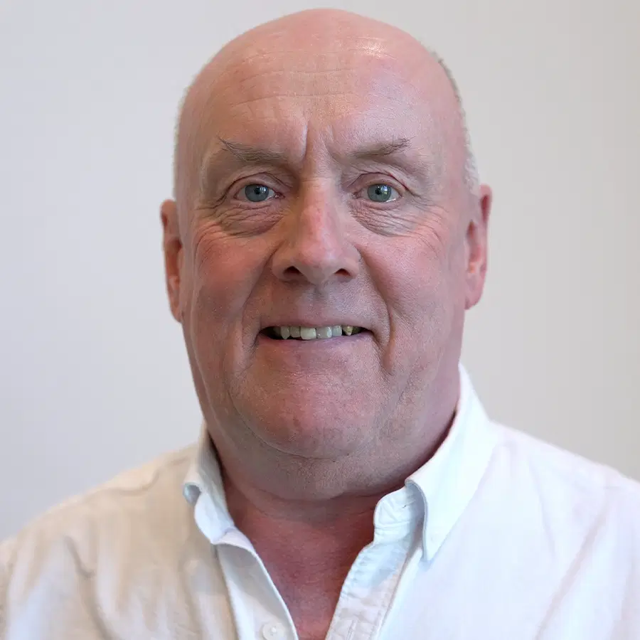 A middle-aged man with a bald head and light skin smiles at the camera, wearing a white button-up shirt, against a plain light background.