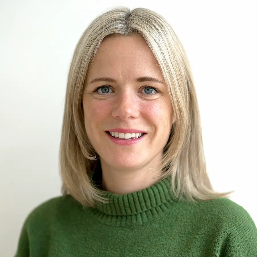 A woman with straight, shoulder-length blonde hair wearing a green jumper smiles at the camera against a plain white background.