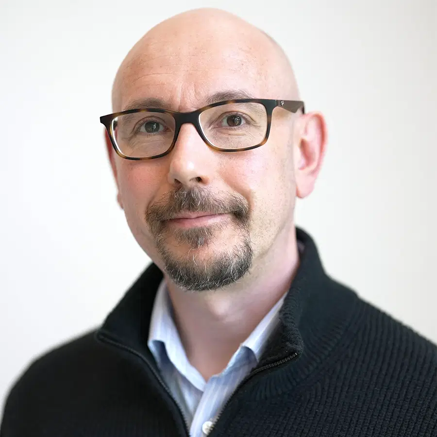 A man with glasses, a trimmed goatee, and a bald head, wearing a dark zip-up jumper over a collared shirt, poses against a plain light background.
