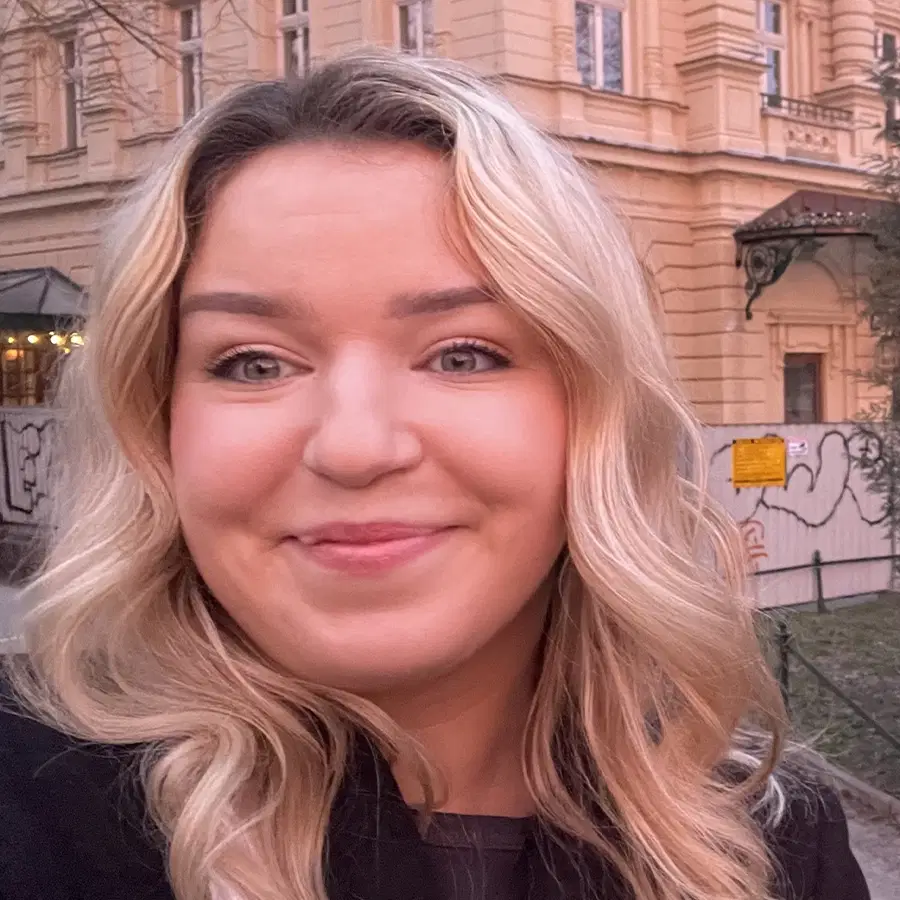 A woman with long, wavy blonde hair smiles in front of a historic yellow building with decorative details and trees in the background.