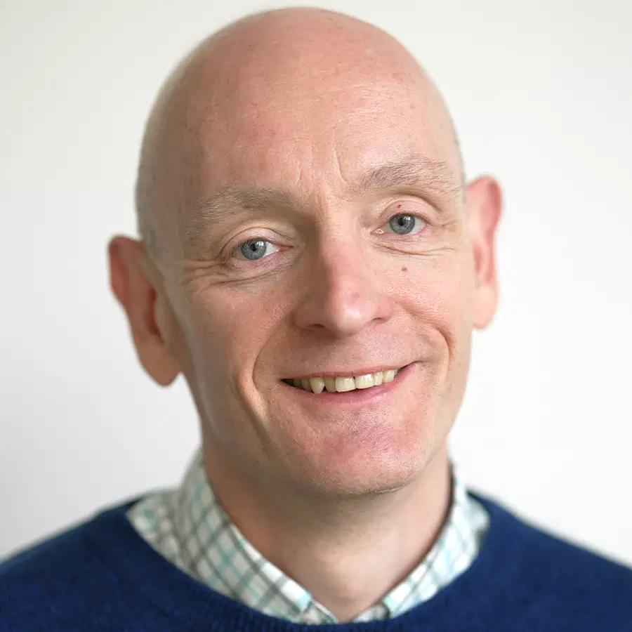 A smiling bald man in a blue jumper over a chequered shirt poses in front of a plain light background.