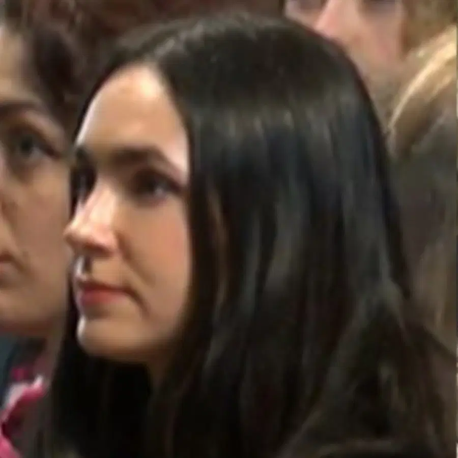 A woman with long dark hair and light skin looks ahead with a neutral expression. Another person is partially visible beside her. The background is out of focus and crowded.