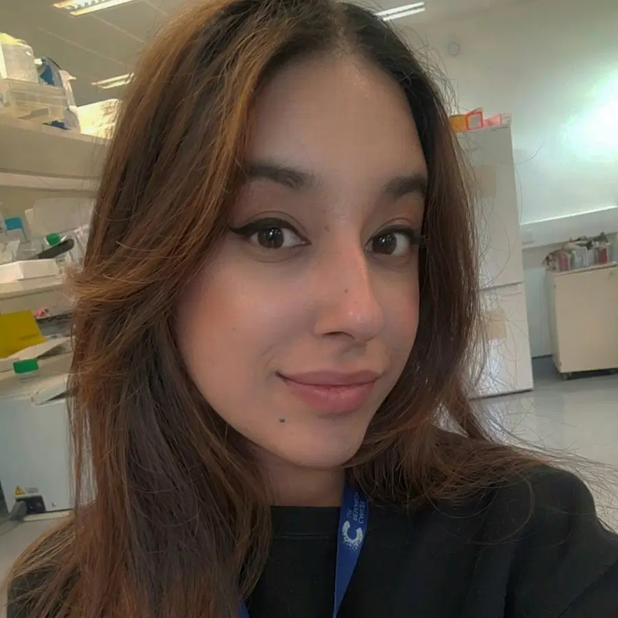 A person with long brown hair and a blue lanyard smiles slightly whilst taking a selfie in a bright, modern laboratory setting. Shelves, equipment, and white cabinetry are visible in the background.