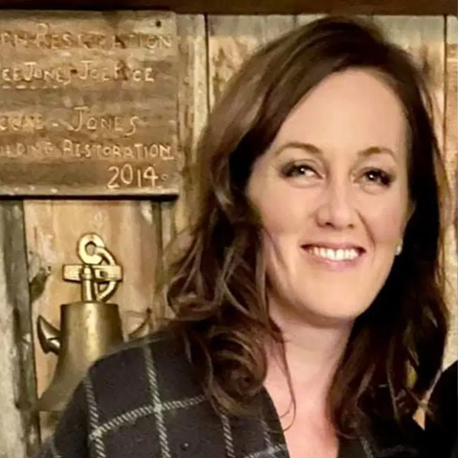 A woman with wavy brown hair smiles at the camera. She stands in front of a wooden wall with plaques and a brass bell. The image is warmly lit and the woman wears a checked-patterned jacket.