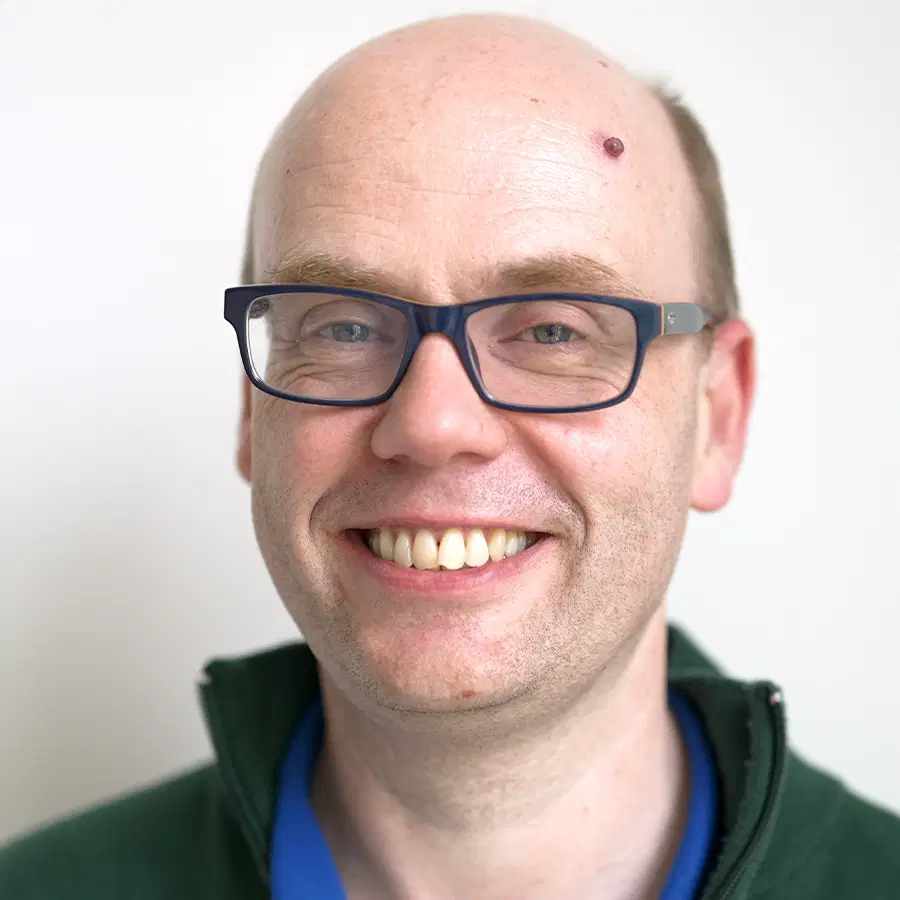 A smiling man with glasses and a mole on his forehead, wearing a green jacket over a blue shirt, standing in front of a plain white background.