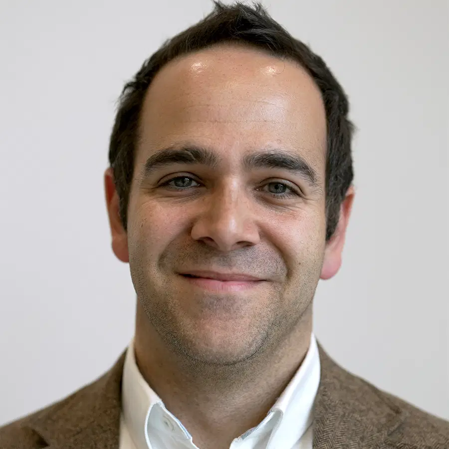 A man with short dark hair, wearing a brown jacket and white shirt, smiling at the camera against a plain light background.