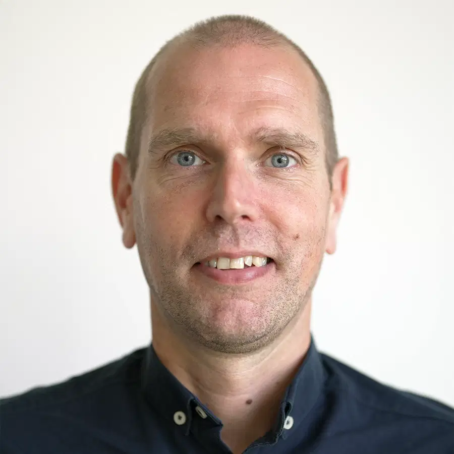 A man with short hair, blue eyes, and a slight smile wears a dark buttoned shirt. He is facing forwards against a plain, light-coloured background.
