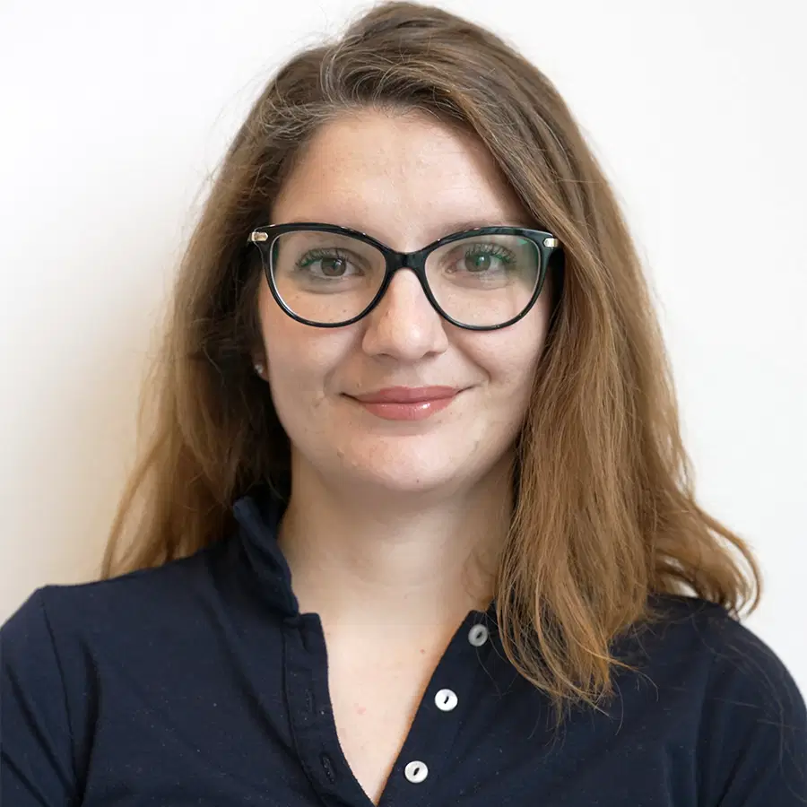 A woman with long brown hair and glasses smiles gently at the camera. She is wearing a navy blue collared shirt and stands against a plain white background.