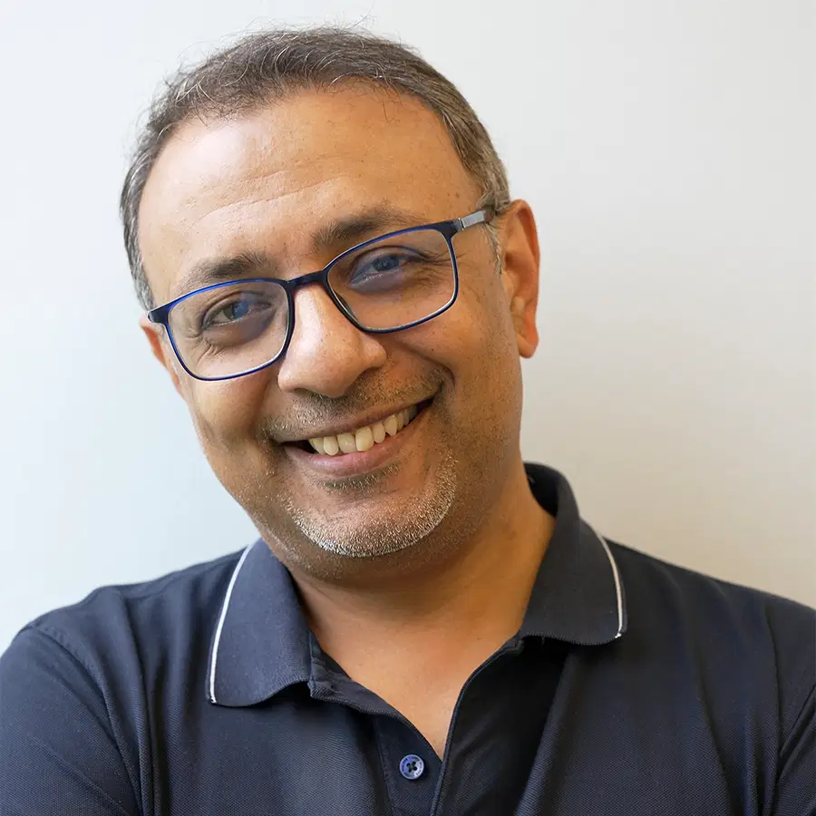 A middle-aged man with short hair, glasses, and a beard is smiling. He is wearing a navy blue polo shirt and standing against a plain light background.