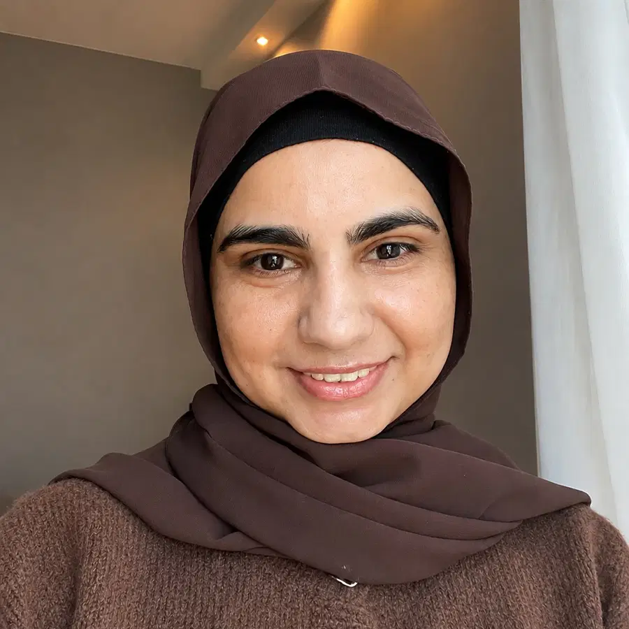 A woman wearing a brown hijab and brown jumper smiles at the camera, standing indoors near a window with soft natural light.