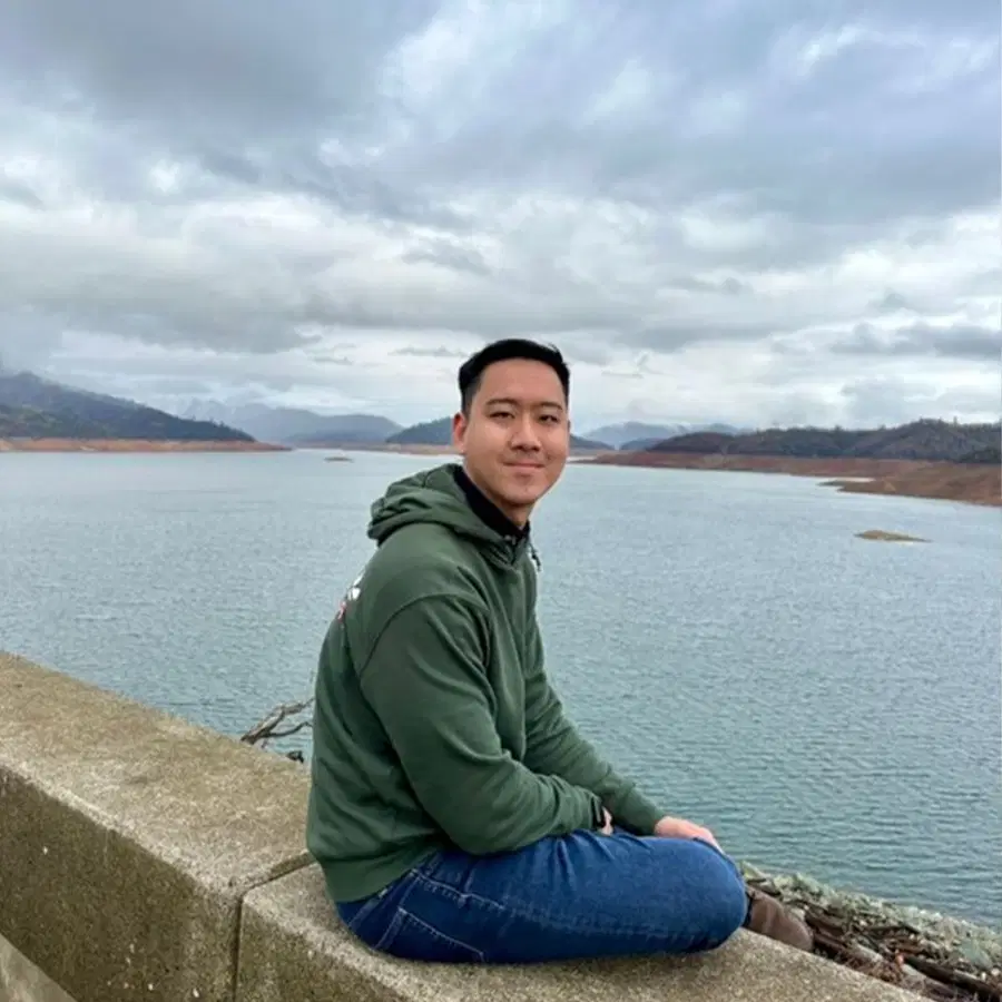 A person in a green hoodie and jeans sits on a concrete ledge by a large lake with mountains and cloudy skies in the background.