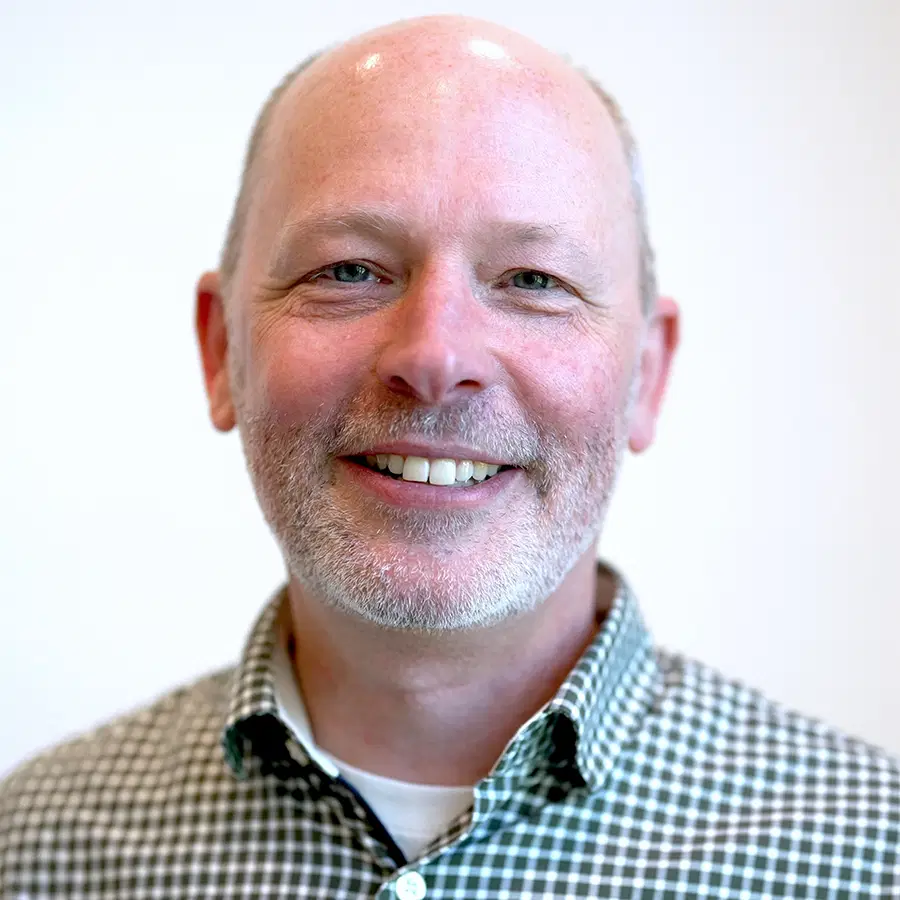 A middle-aged man with a bald head and short beard, smiling. He is wearing a black and white chequered shirt with a white T-shirt underneath, with a plain light background.