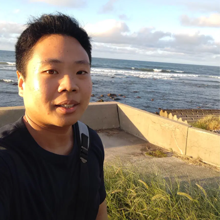 A person takes a selfie near the sea, standing by a low wall. Waves crash in the background under a partly cloudy sky, with grass and concrete visible in the foreground.