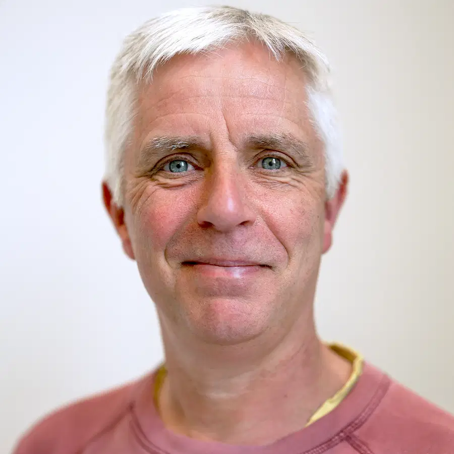 A middle-aged man with short grey hair and blue eyes is smiling softly at the camera. He is wearing a faded red jumper and is posed against a plain, light background.