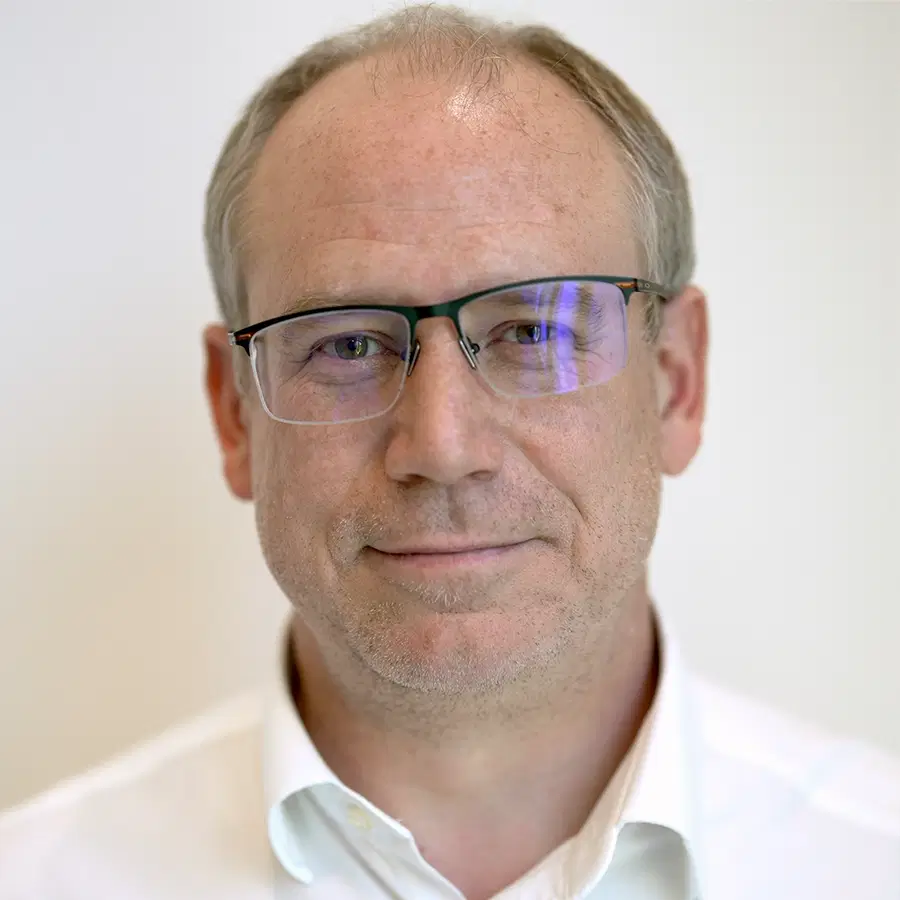 Middle-aged man with short fair hair, glasses, and a light-coloured collared shirt, standing against a plain white background, looking directly at the camera with a neutral expression.