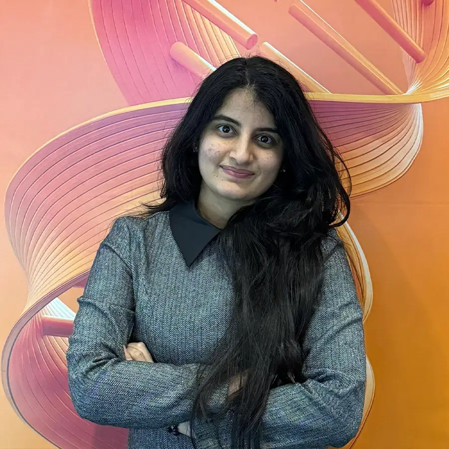 A woman with long dark hair, wearing a grey dress with a black collar, stands smiling with arms crossed in front of an orange background featuring a stylised DNA double helix design.