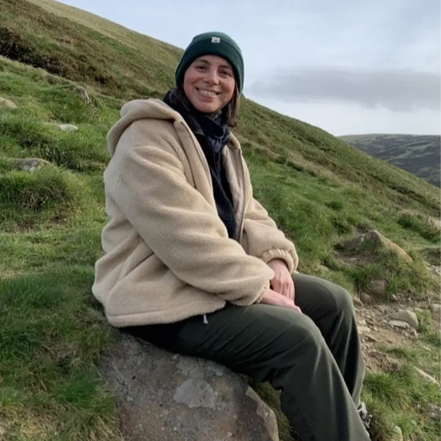 A person in a beige jacket and black beanie sits on a rock, smiling, surrounded by grassy hills under a cloudy sky.
