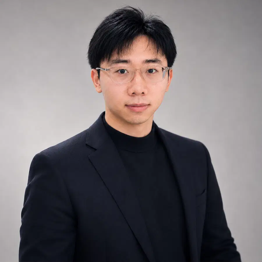 A man wearing glasses and a black suit with a black shirt stands against a plain grey background, facing the camera with a neutral expression.