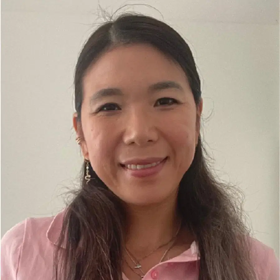 A woman with long dark hair, wearing a pink collared shirt and jewellery, smiles at the camera in a well-lit room with a plain light-coloured wall in the background.