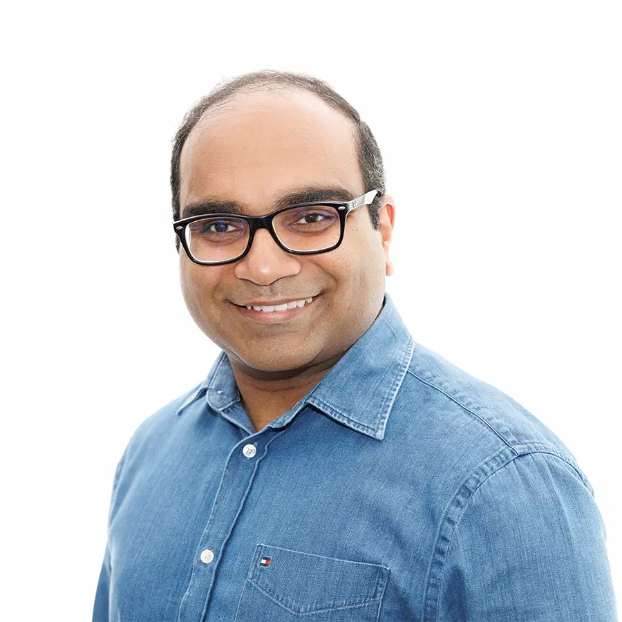 A smiling man with glasses and short hair wears a blue denim button-up shirt, standing against a plain white background.