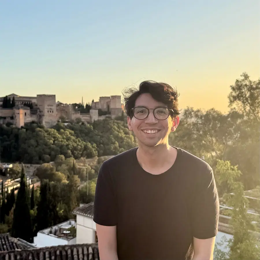 A person with glasses and dark hair smiles outdoors at sunset, with historic buildings, trees, and a scenic landscape in the background.