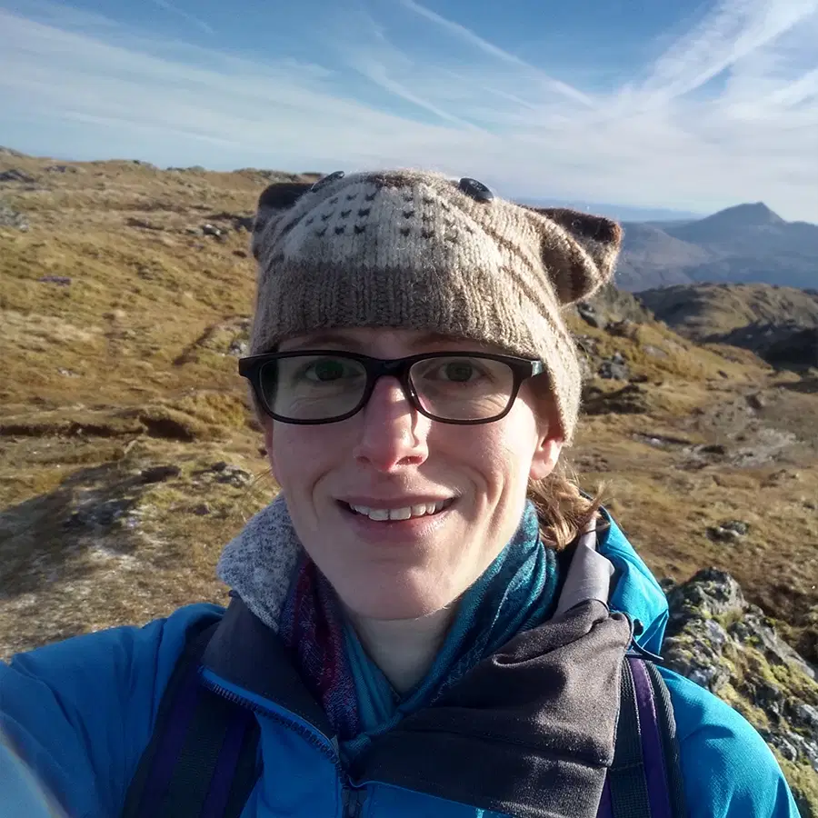 A person wearing glasses, a blue jacket, and a knitted hat with animal ears smiles outdoors on a rocky, sunlit landscape with mountains in the background.