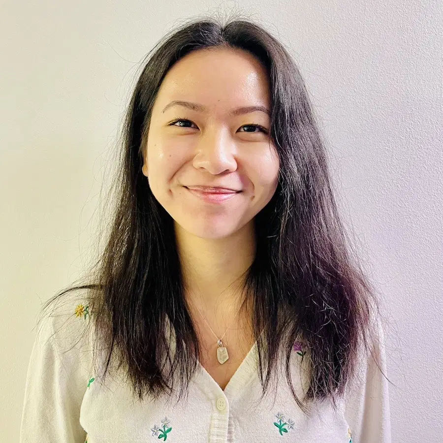 A woman with long dark hair smiles at the camera, wearing a light-coloured shirt with small embroidered designs and a pendant necklace, standing against a plain light-coloured wall.