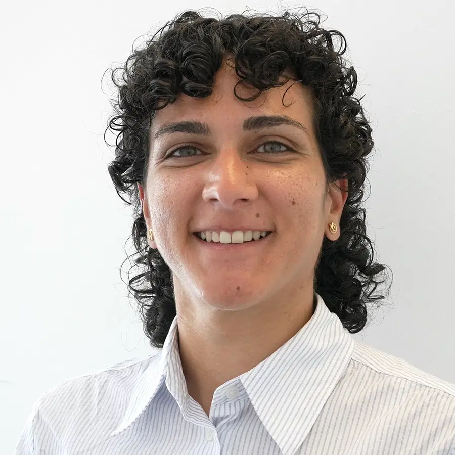 Person with short, curly dark hair and light skin smiling, wearing a light striped button-up shirt, standing against a plain white background.